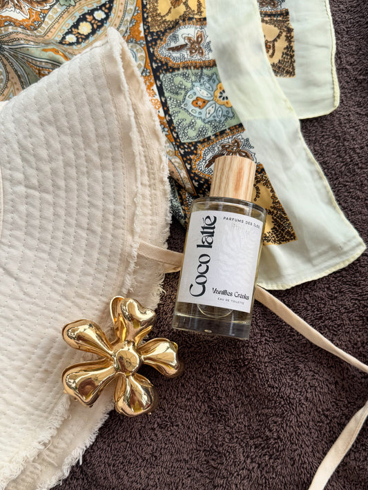 eau de Toilette COCO LATTE 100ML AUF EINEM HANDTUCH. Daneben ein Sonnenhut und eine Blumenhaarspange