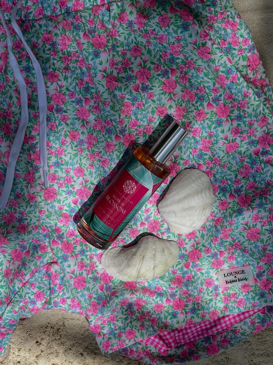 Parfüm Tropical Blossom liegend auf einer Badehose mit Blumenmustern im Sand. Neben dem Parfüm liegen Muscheln