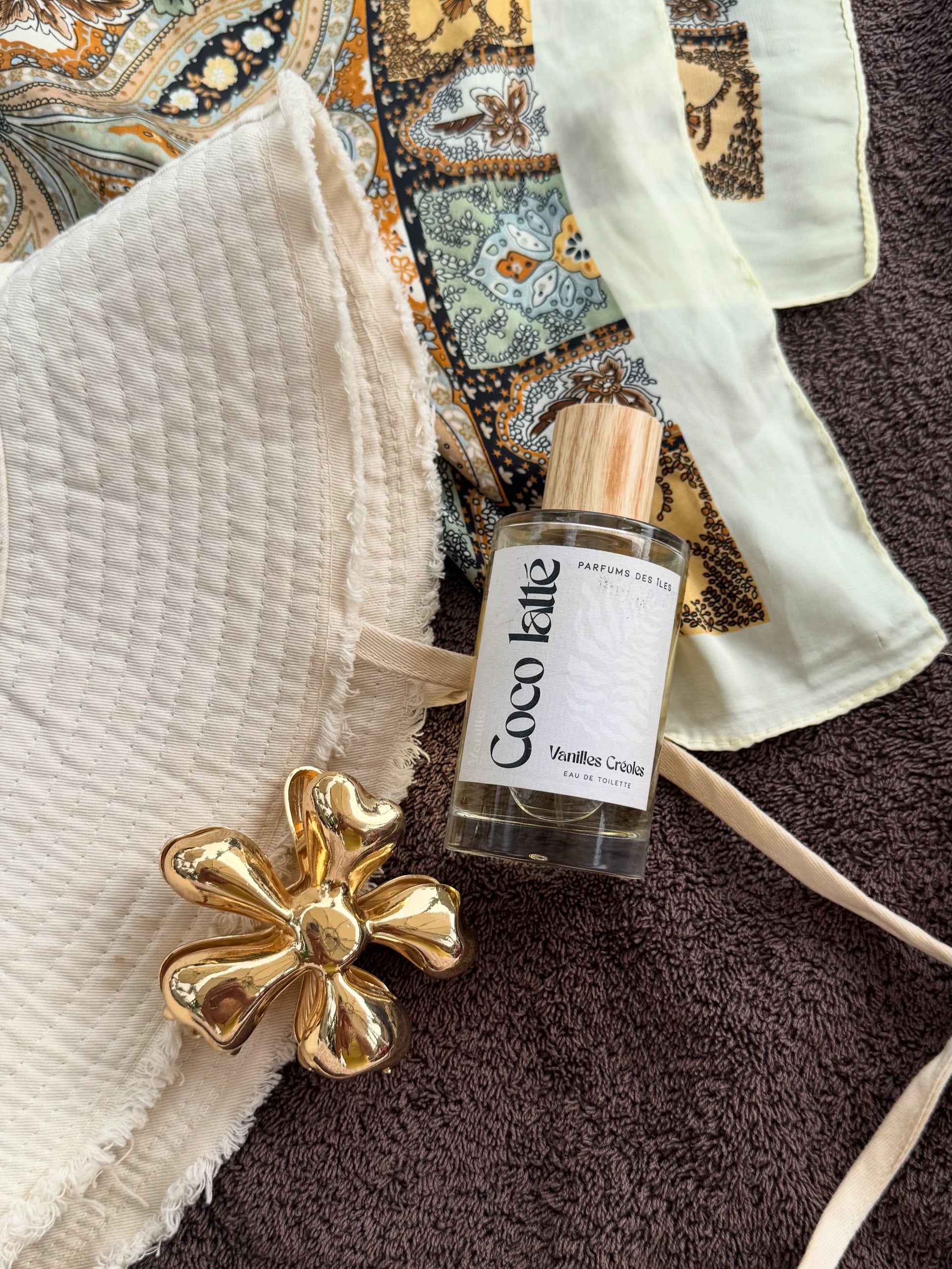eau de Toilette COCO LATTE 100ML AUF EINEM HANDTUCH. Daneben ein Sonnenhut und eine Blumenhaarspange