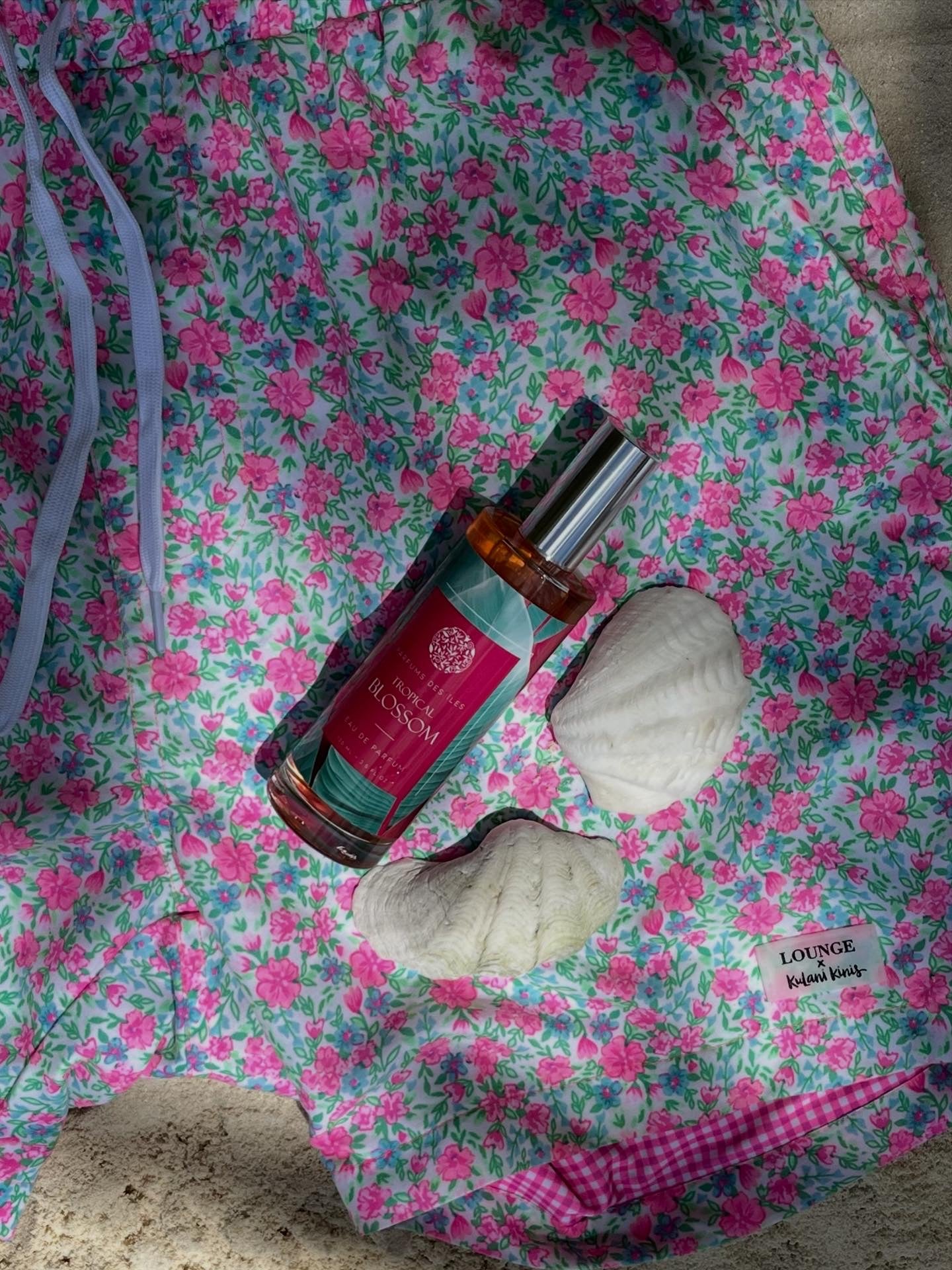 Parfüm Tropical Blossom liegend auf einer Badehose mit Blumenmustern im Sand. Neben dem Parfüm liegen Muscheln