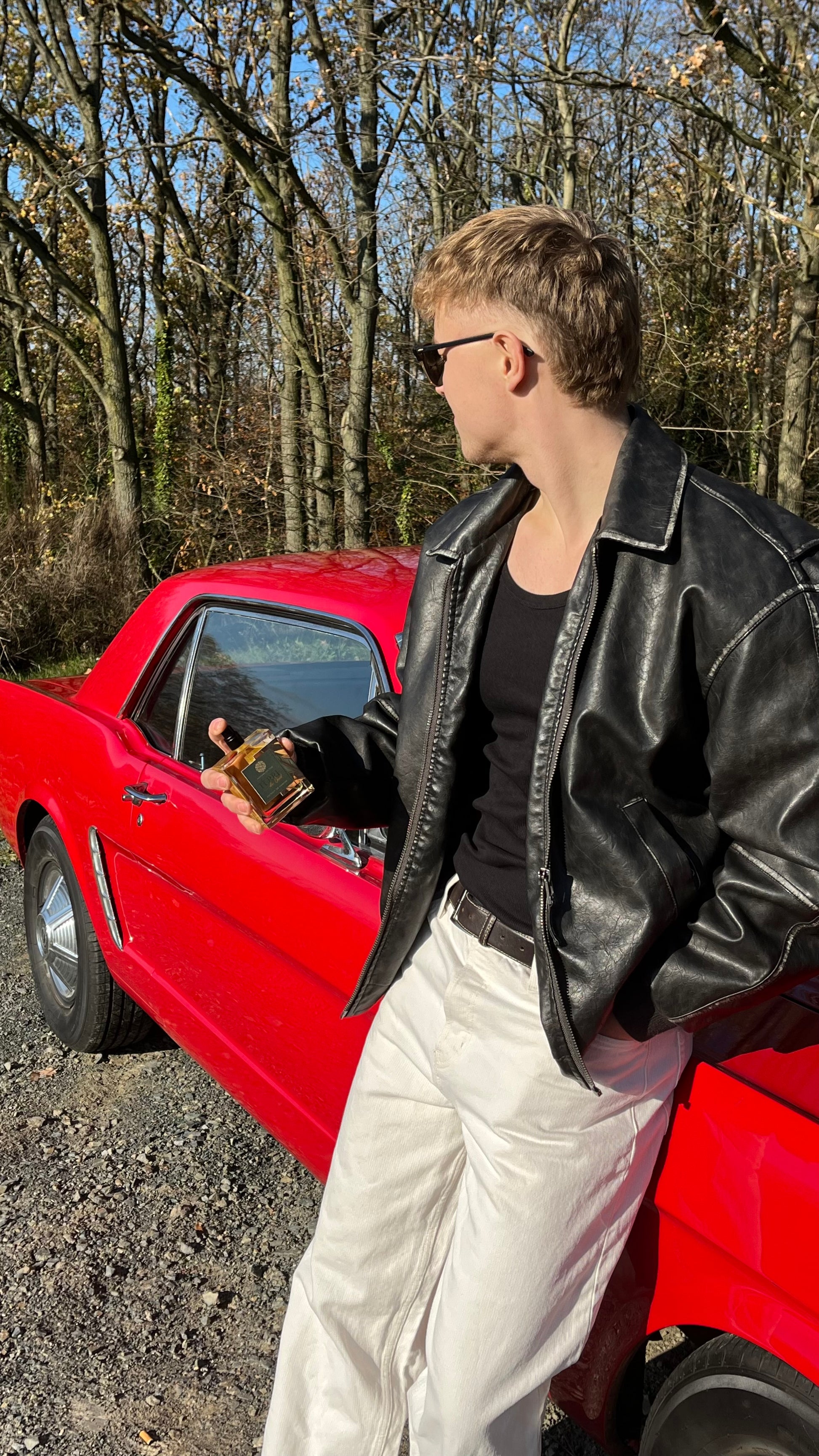 Rotes Auto im Hintergrund. Männliches Model hält das Parfüm Tere de Oud in den Händen. Model trägt weiße Hose mit schwarzer Lederjacke. Im Hintergrund Bäume
