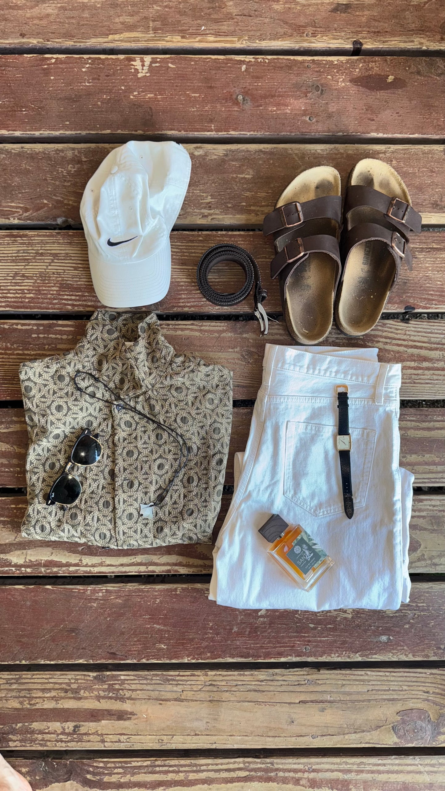 Outfit liegend auf einer Holzterrasse. Bestehend aus Schuhen, Gürtel, Cap, Hemd, Kette, Sonnenbrille und weißer Hose. Auf der Hose liegend das Santal Parfüm