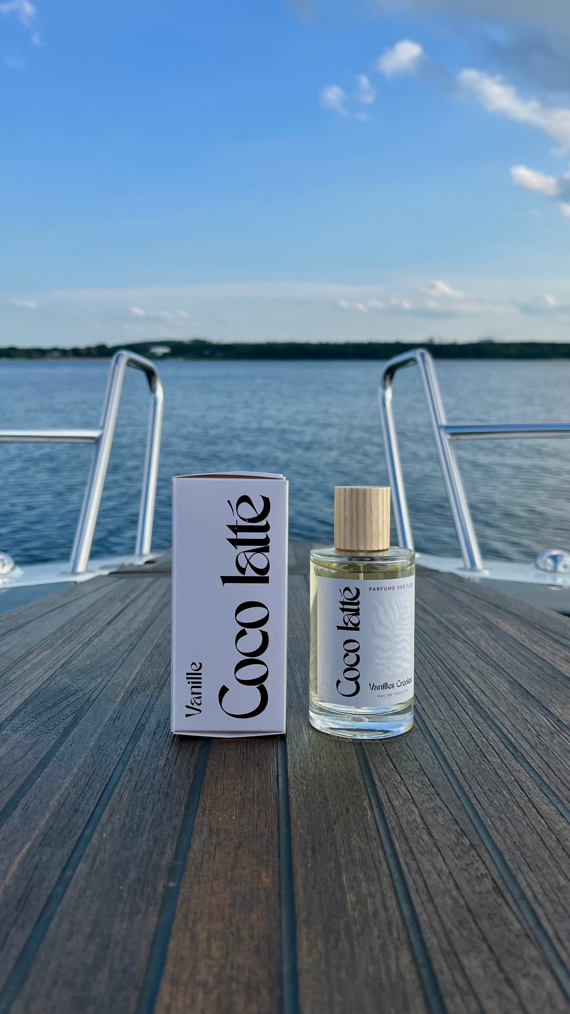 Eau de Toilette Coco Latte 100ml links Karton rechts Flacon. Stehend auf einem Boot im Hintergrund die Reeling und ein See