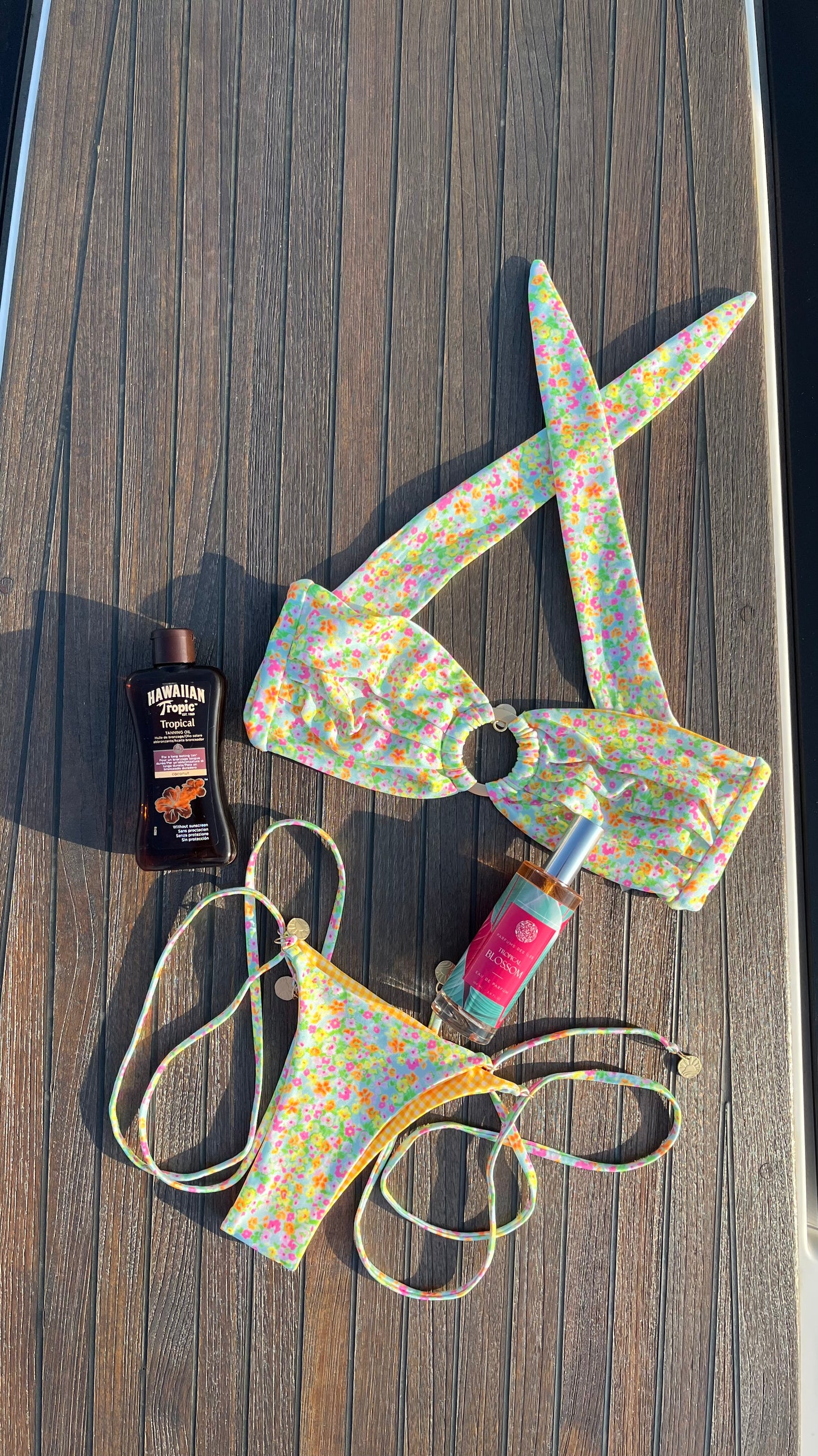 Eau de Parfum Tropical Blossom 100ml Flacon liegend zwischen einem Bunten Bikini auf einem Boot. Links eine Flasche Sonnenschutz