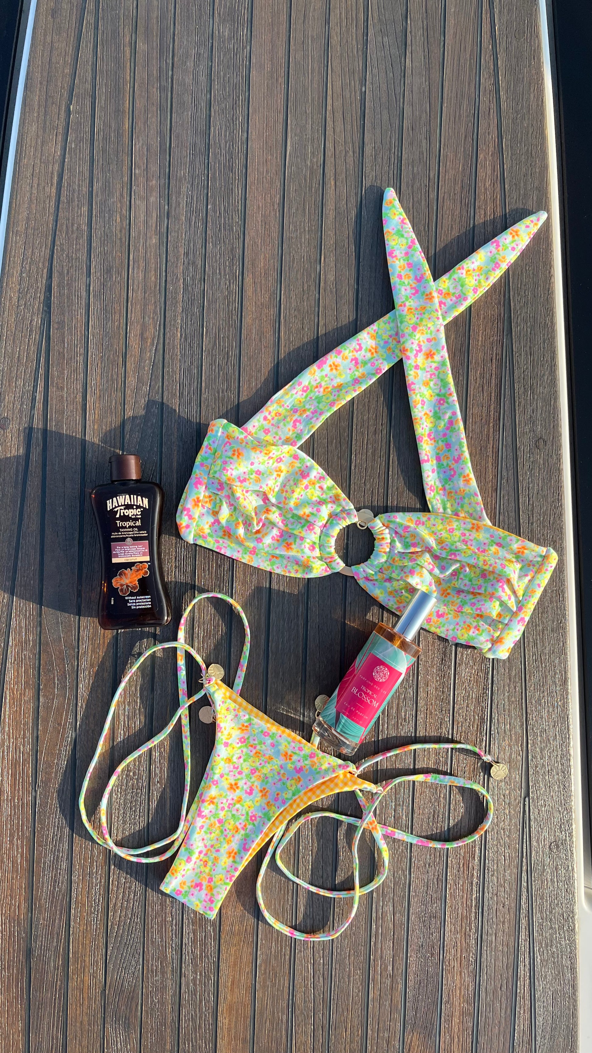 Eau de Parfum Tropical Blossom 100ml Flacon liegend zwischen einem Bunten Bikini auf einem Boot. Links eine Flasche Sonnenschutz