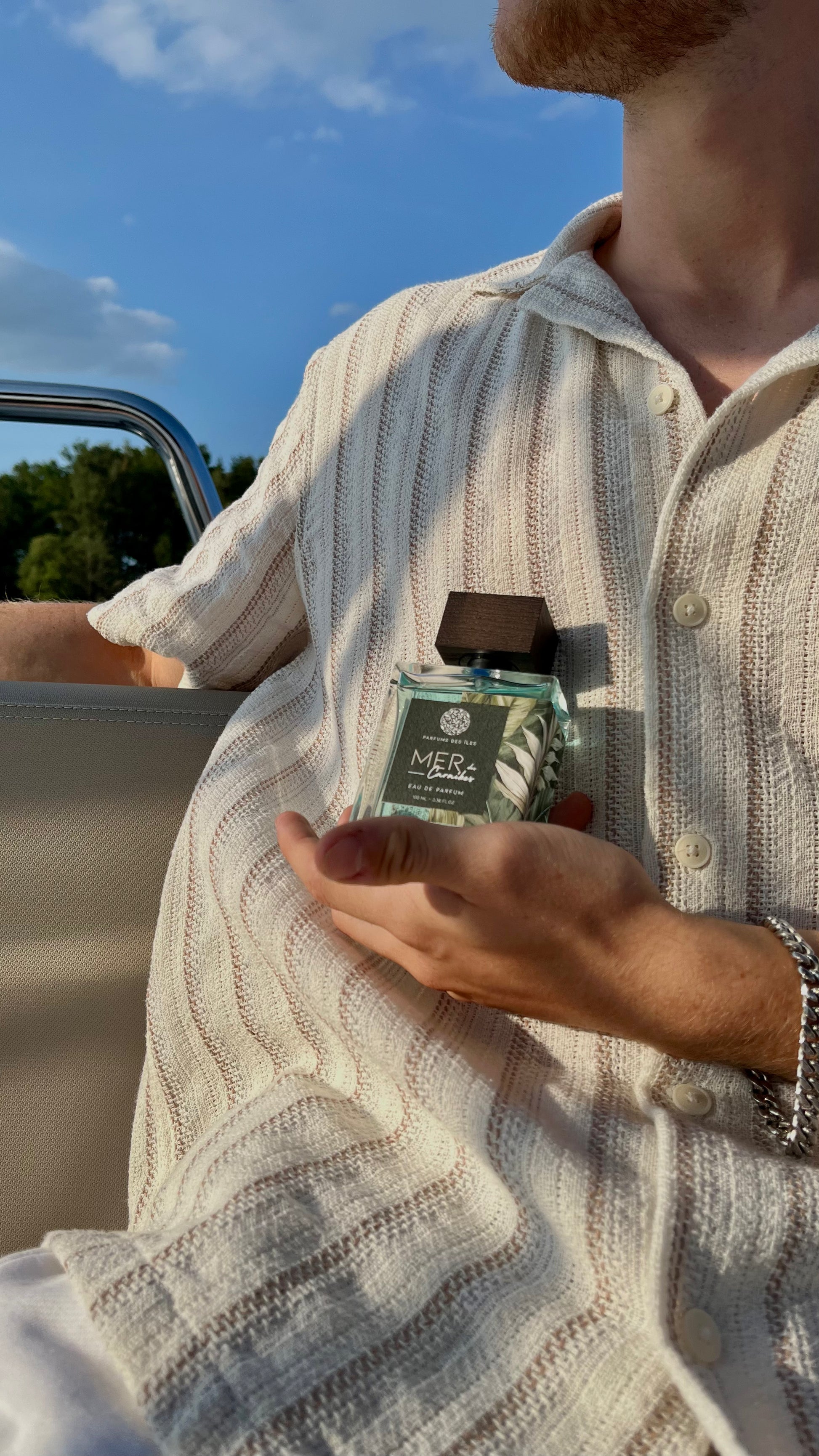 Eau de Parfum Mers des Caraibes 100ml Flacon gehalten in der Hand vor dem Körper. Körper trägt ein Leinenhemd. Links ist die Reeling eines Bootes.