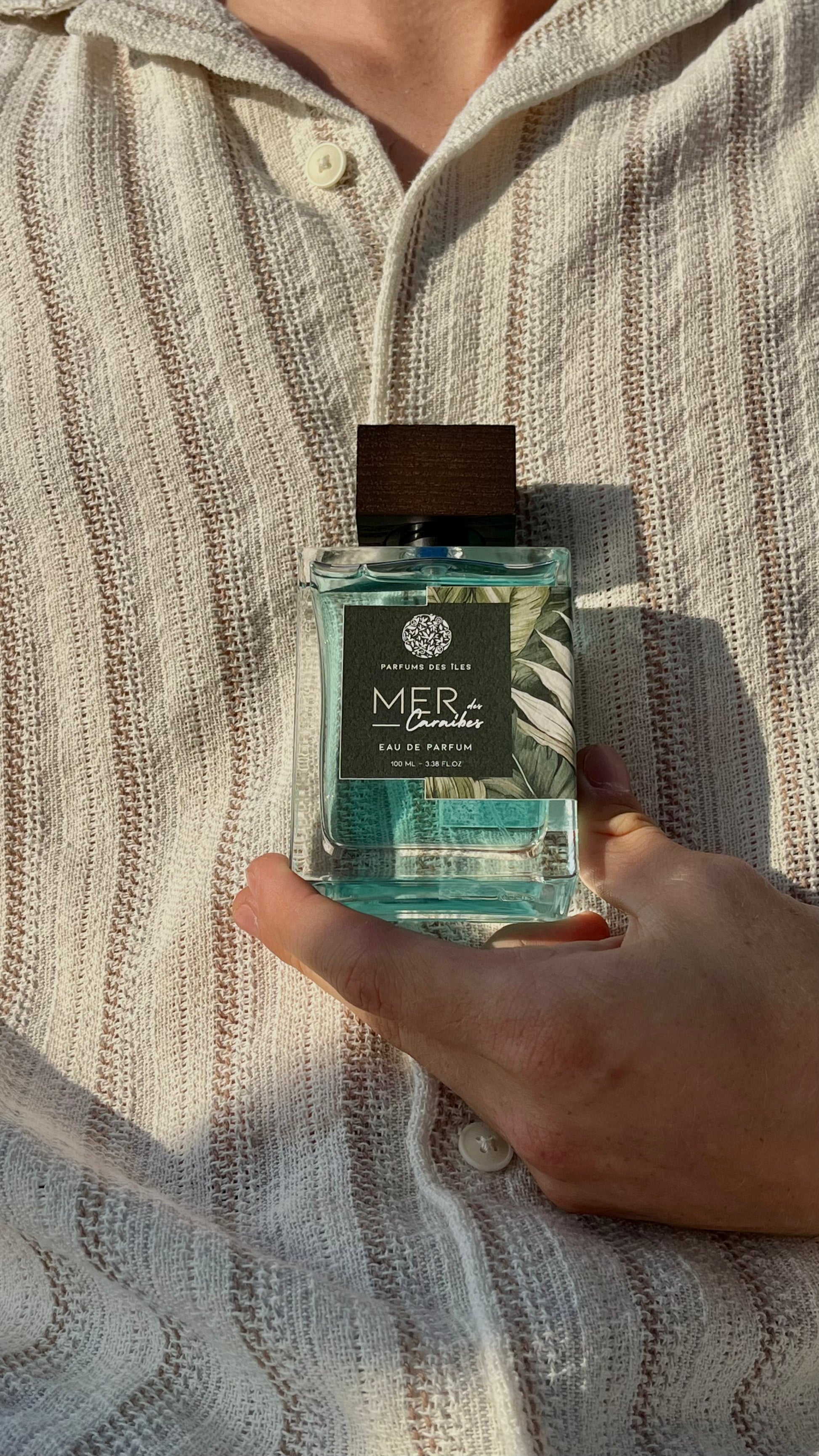 Eau de Parfum Mers des Caraibes 100ml Flacon gehalten in der Hand vor dem Körper. Körper trägt ein Leinenhemd