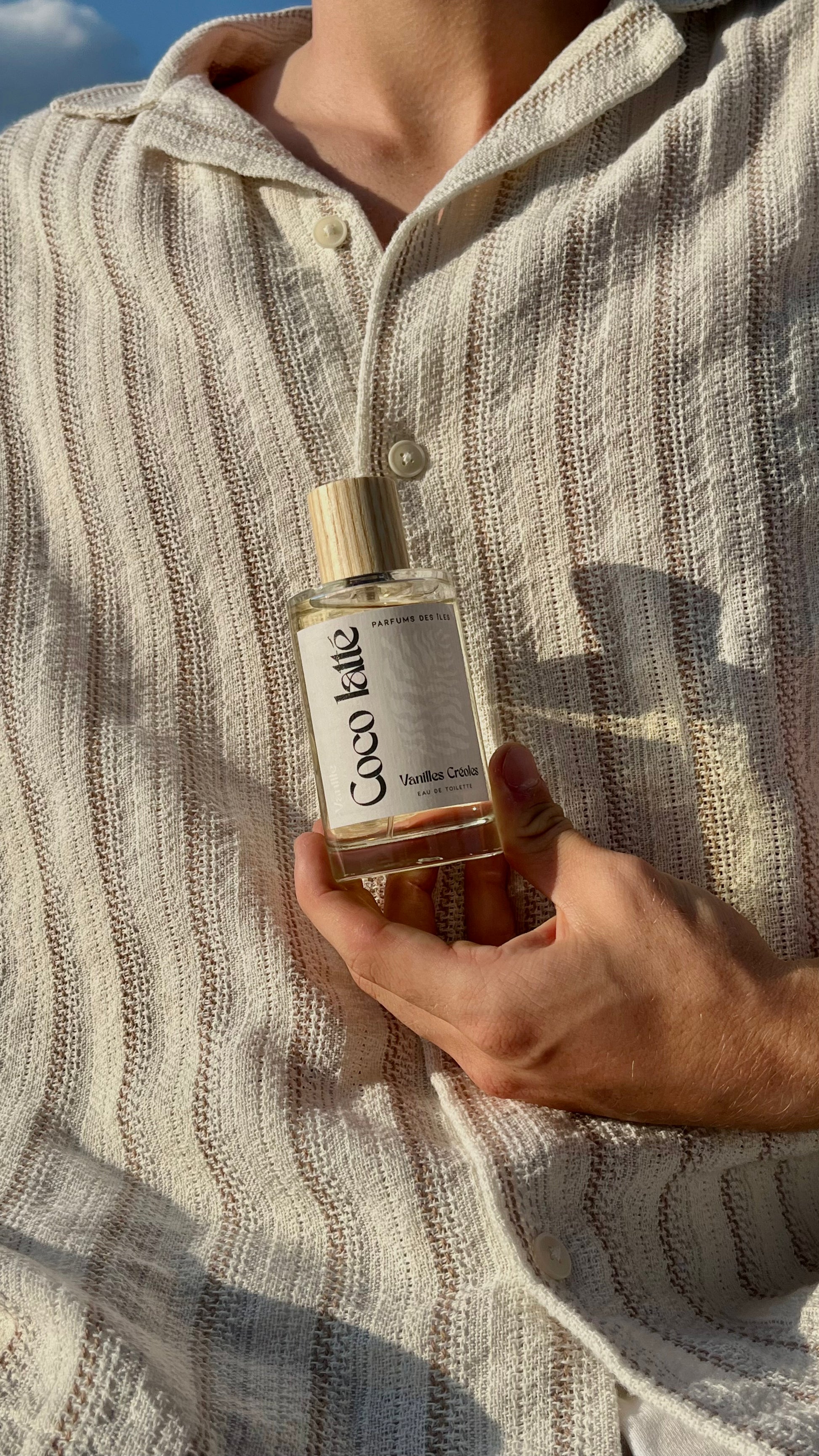 Eau de Toilette Coco latte 100ml. Gehalten in der Hand vor dem Körper. Der Körper trägt ein Leinenhemd.