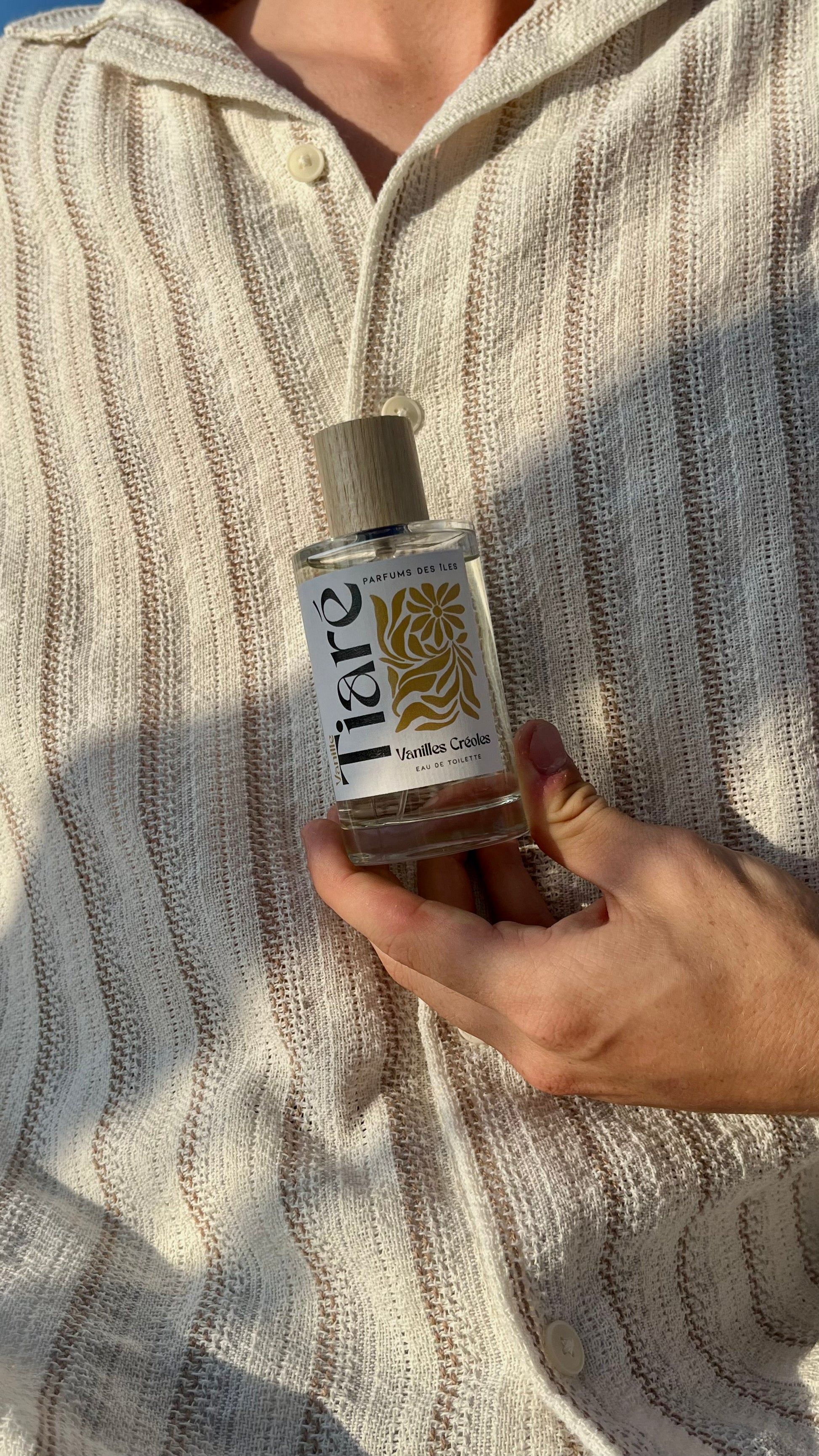 Eau de Toilette Tiare 100ml. Gehalten in der Hand vor dem Körper. Der Körper trägt ein Leinenhemd.