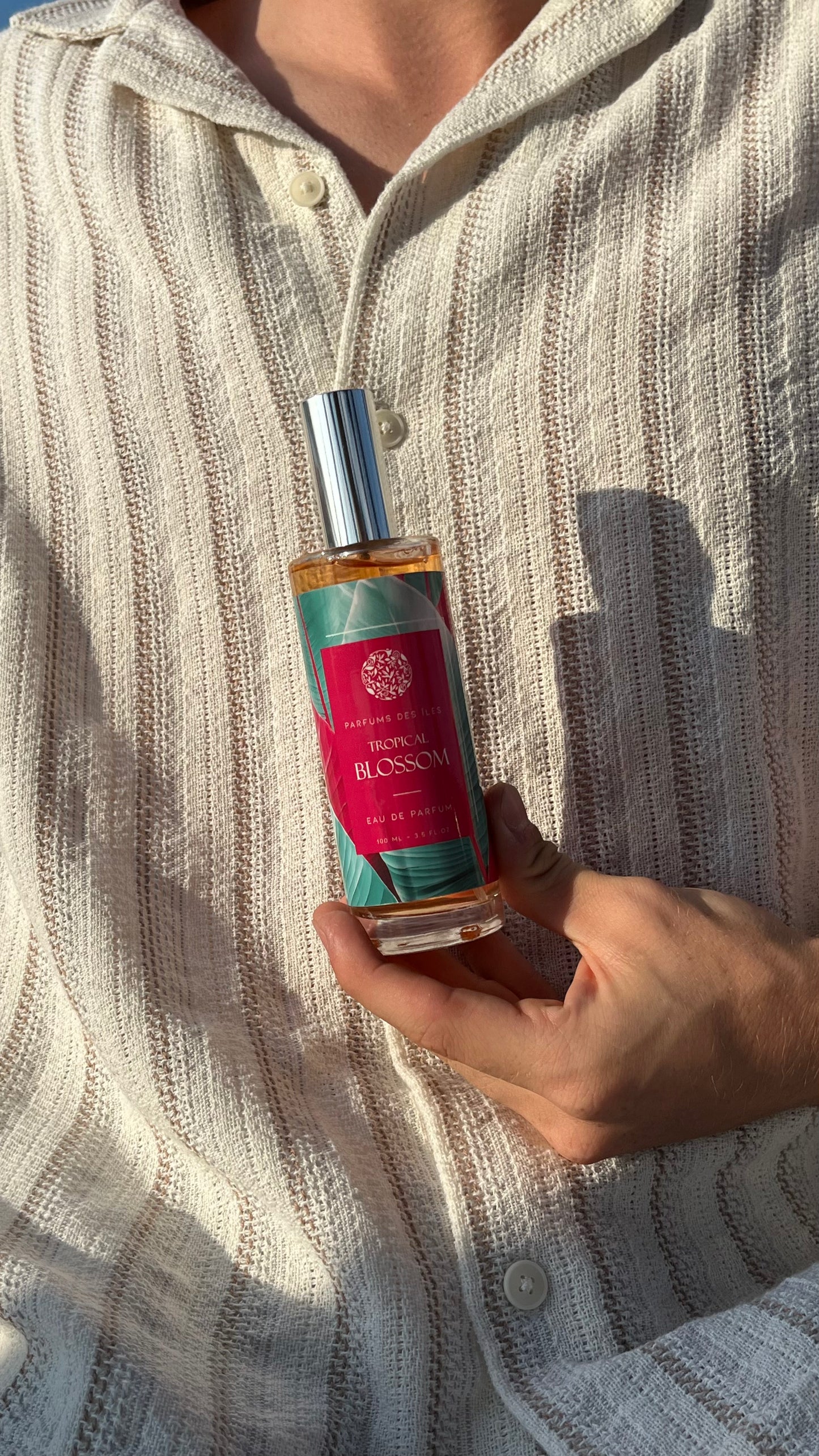 Tropical Blossom Parfüm 100ml. Gehalten in der Hand vor dem Körper. Der Körper trägt ein Leinenhemd.