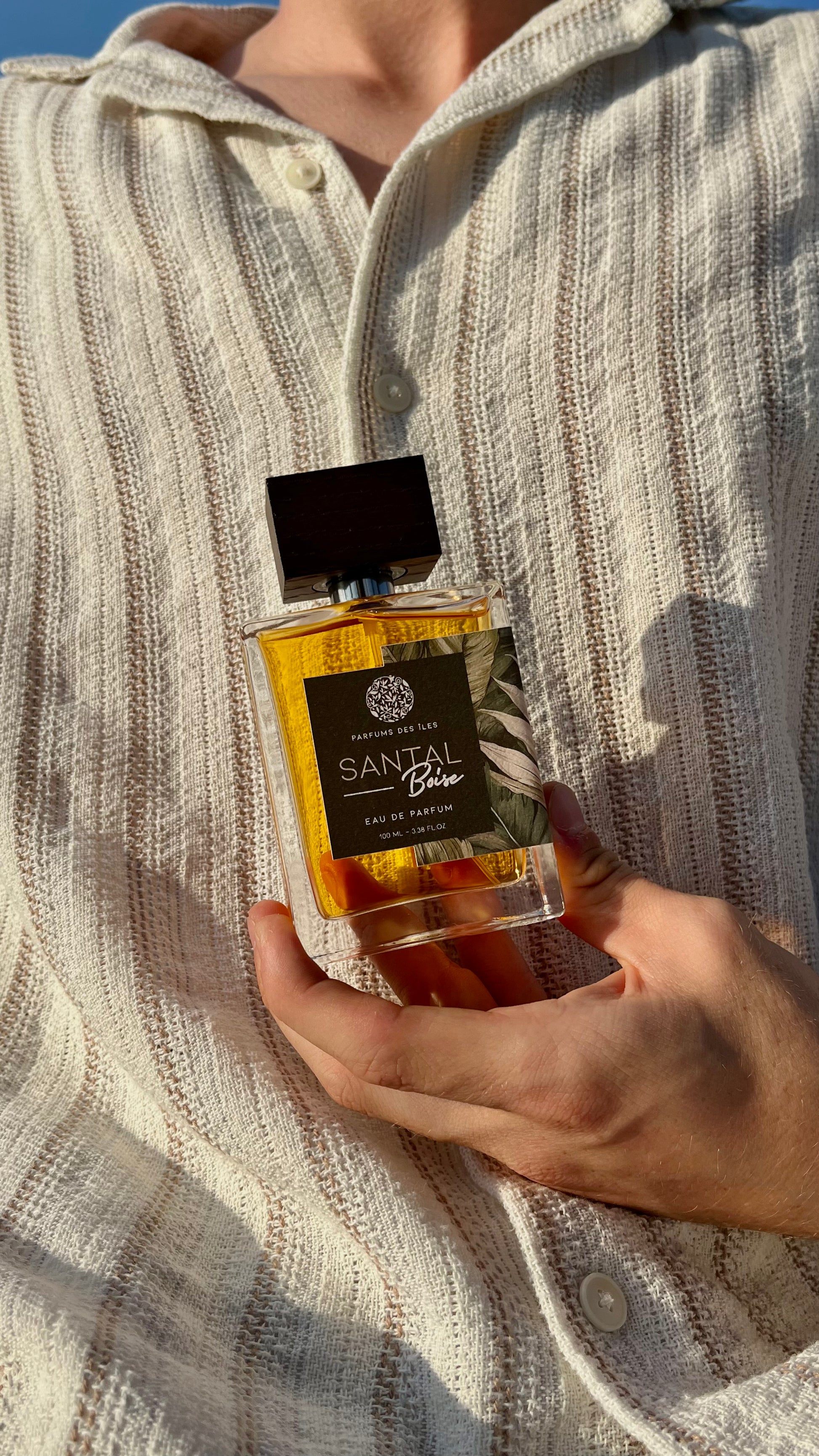 Eau de Parfum Santal 100ml in der Hand vor dem Körper gehalten. Körper trägt ein Leinenhemd