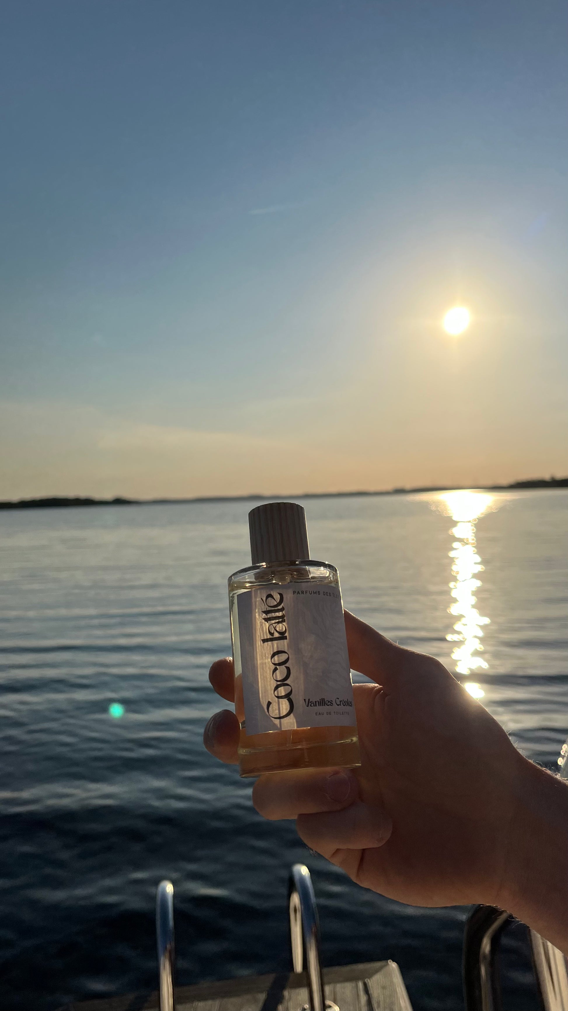 Eau de Toilette Coco Latte. 100ml in der Handgehalten. Im Hintergrund Bootstreppe und ein See im Sonnenuntergang.