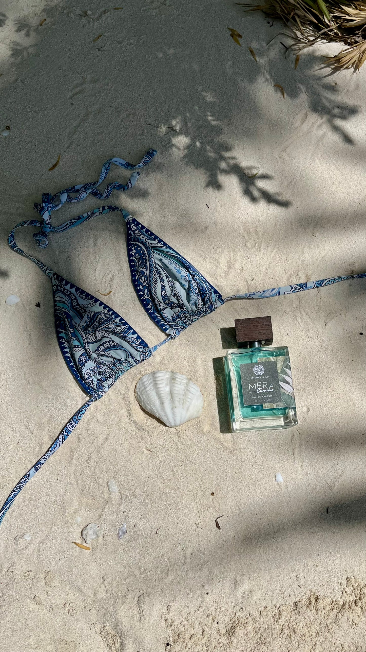 Parfüm Mer des Caraibes liegen im Sand neben einer Muschel und einem Biknioberteil