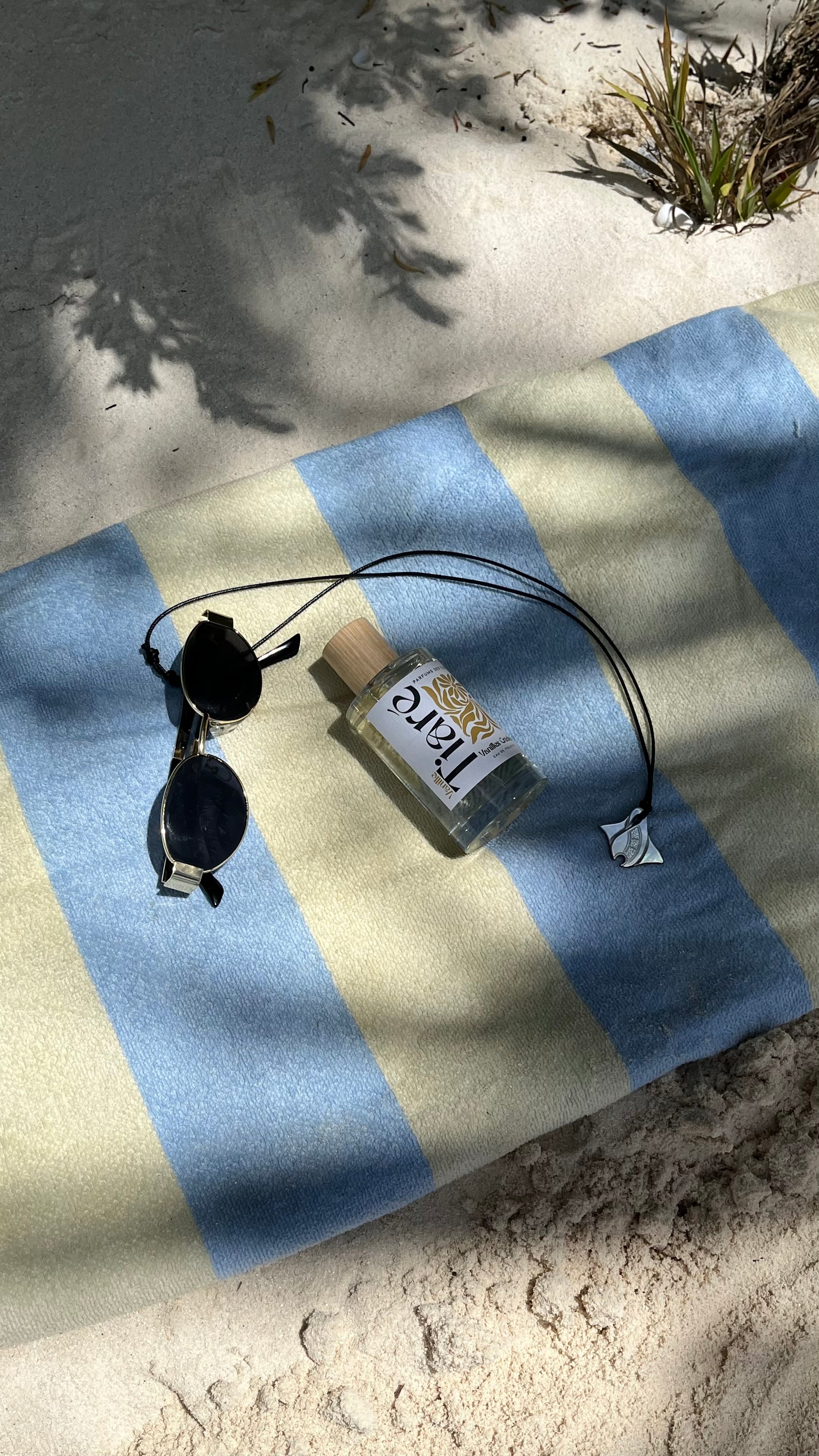 Eau de Toilette Tiaré. Liegend auf einem Handtuch im Sand. Sonnenbrille und Kette daneben.